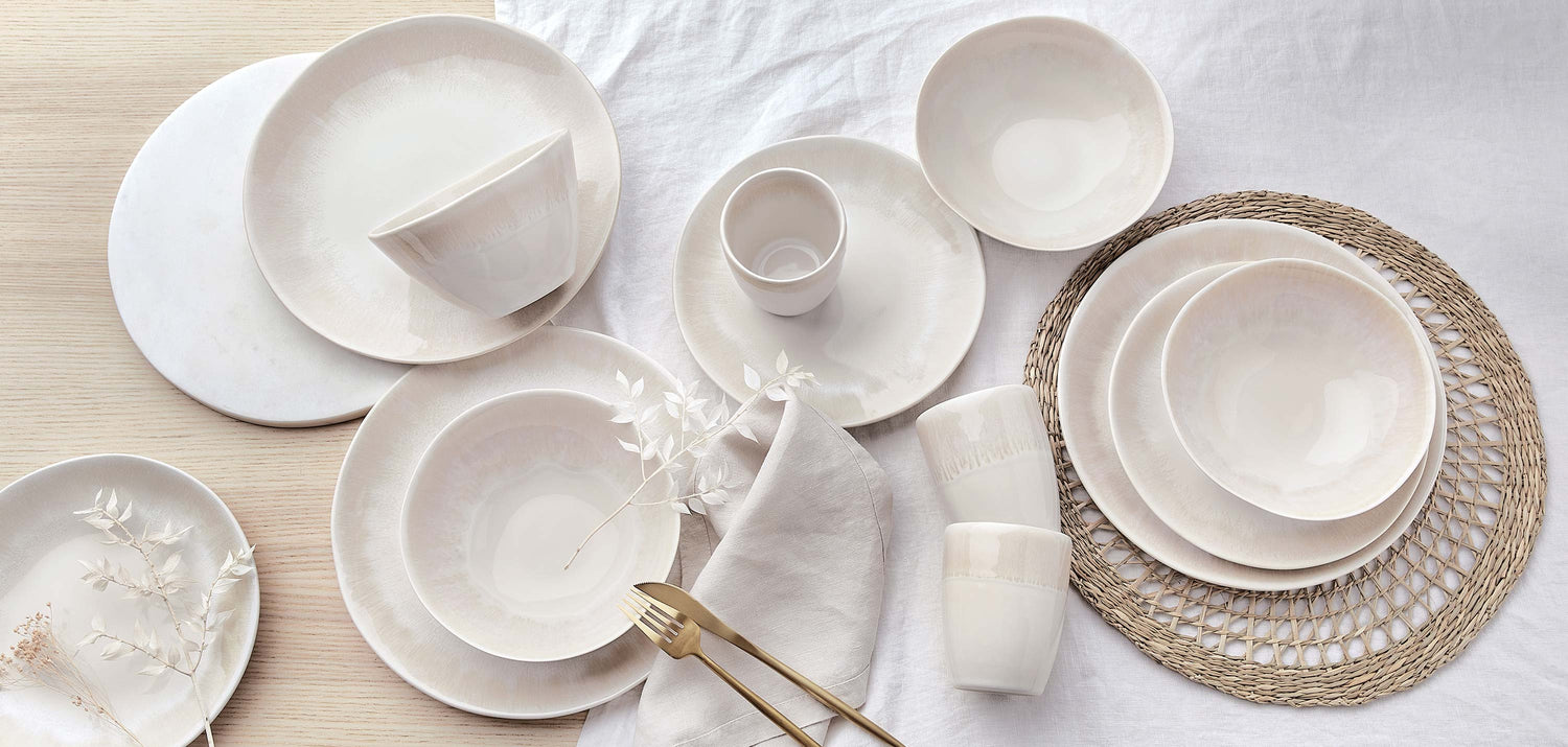 Cremeweißes Steinzeug-Geschirrset mit Tellern, Schüsseln und Bechern auf einem Jute-Tischset und weißem Leinentuch im minimalistischen Scandi-Stil.