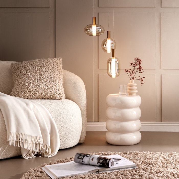 Wohnzimmer-Deko in Beige: Cremeweißer Beistelltisch mit Vase und Trockenblumen neben einem Sofa mit kuscheligen Wohnaccessoires und dekorativen Hängelampen.