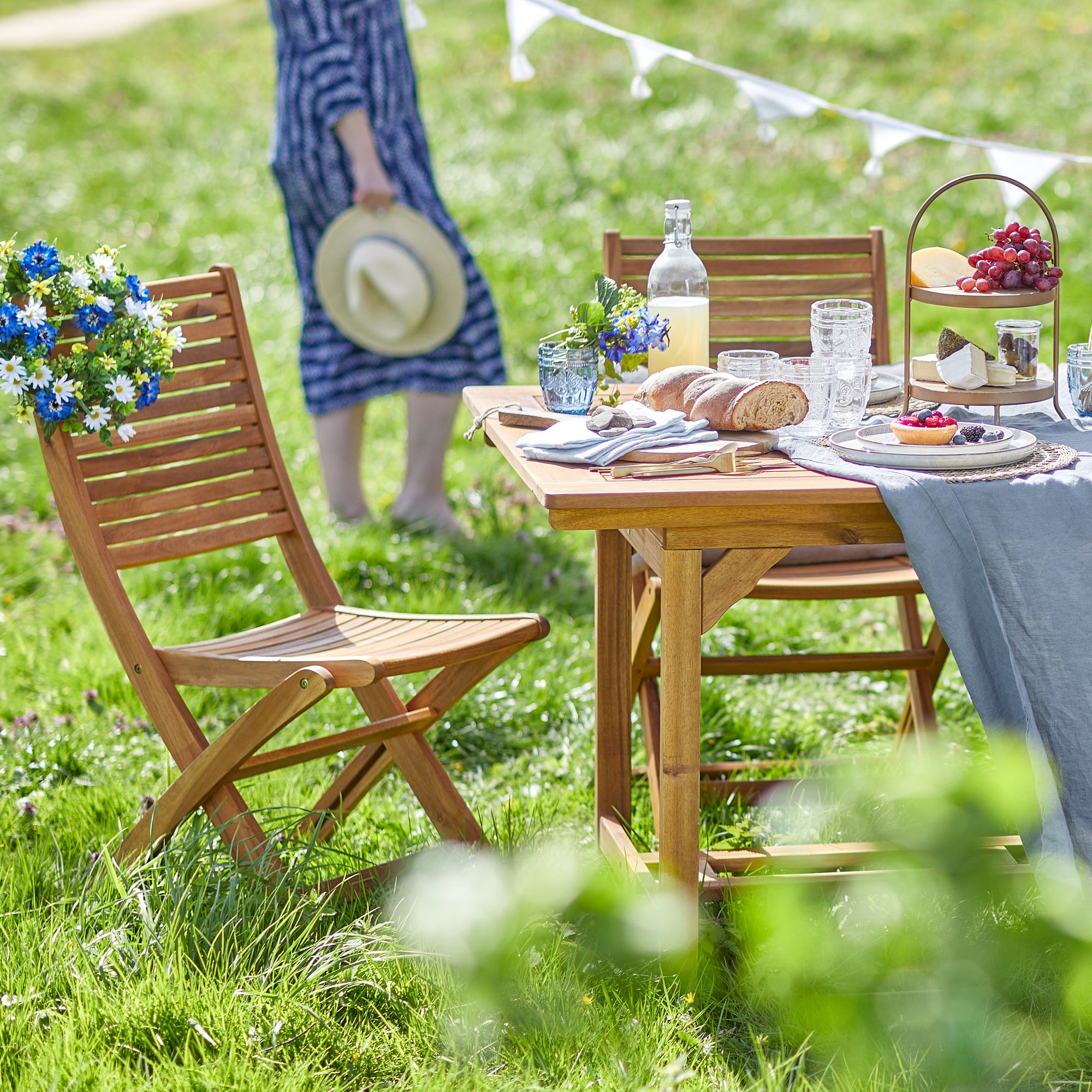 Gedeckter Gartentisch aus Holz mit passenden Klappstühlen auf einer grünen Wiese, dekoriert mit blauem Tischläufer und sommerlichem Frühstück.