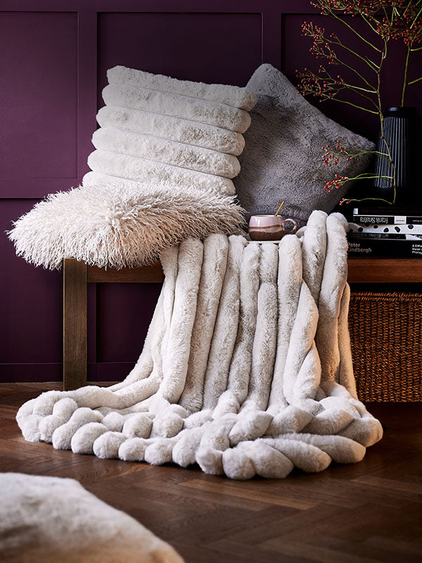 Cremefarbene Kunstfell-Kuscheldecke auf Holzbank, dazu Kissen in Weiß und Grau vor aubergine Wandvertäfelung