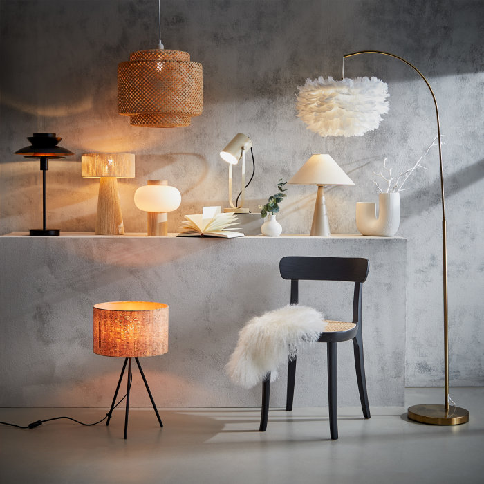 Eine harmonische Auswahl verschiedener Lampen in sanftem Beige und Weiß, darunter filigrane Stehlampen, moderne Hängelampen und gemütliche Tischlampen, stilvoll arrangiert vor einer minimalistischen grauen Wand.