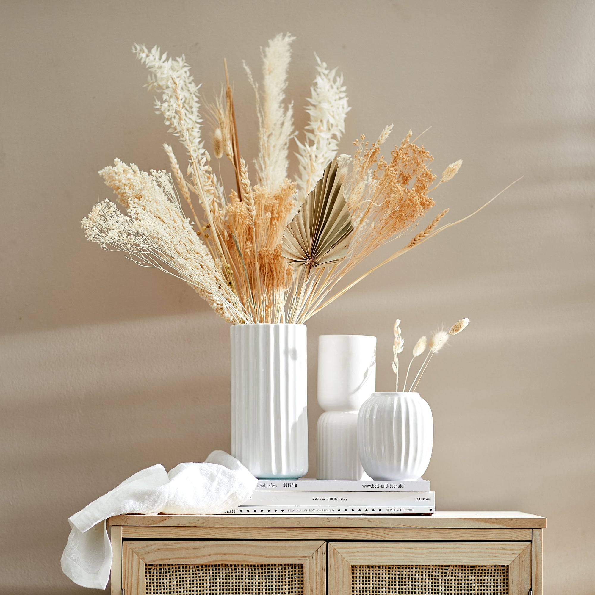 Drei weiße Vasen mit Trockenblumen; unterschiedliche Größen und Formen, arrangiert auf drei Büchern vor einer beigen Wand.