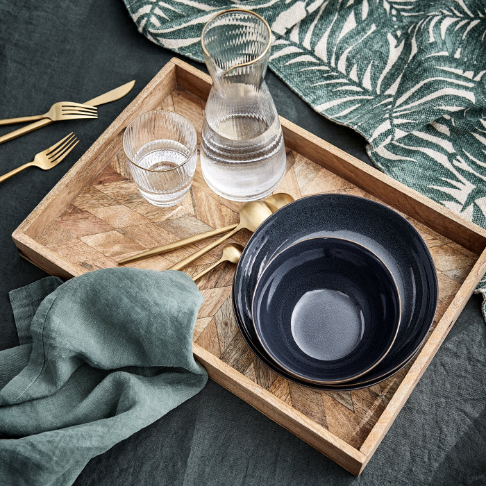 Längliches Tablett aus Holz mit Geschirr, einer Karaffe und Wasserglas, dekorativ platziert auf grau-grünen Laken.