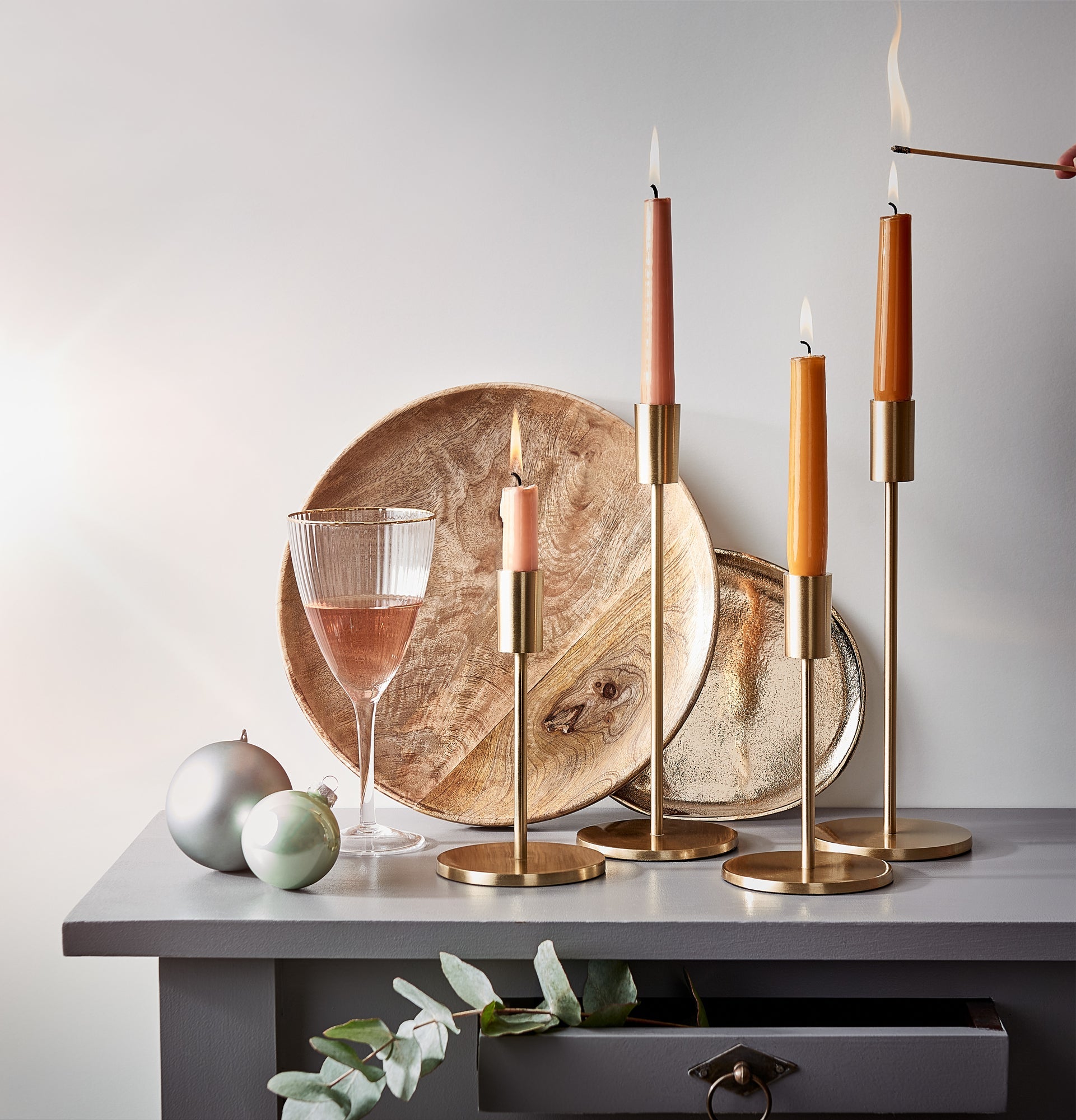 Edle goldene Kerzenhalter für Stabkerzen in Orange und Rosa, stilvoll arrangiert mit goldenen Accessoires auf einem grauen Tisch vor weißer Wand.