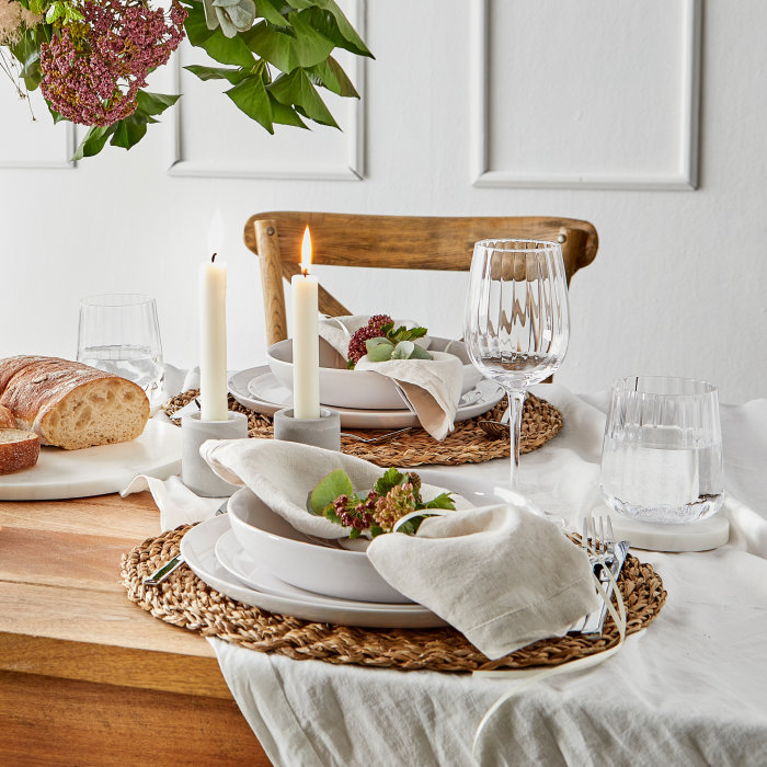 Gedeckter Tisch mit runden Tischsets aus beigem Seegras auf weißer Leinentischdecke, kombiniert mit Besteck und Kerzen auf einem Holztisch.