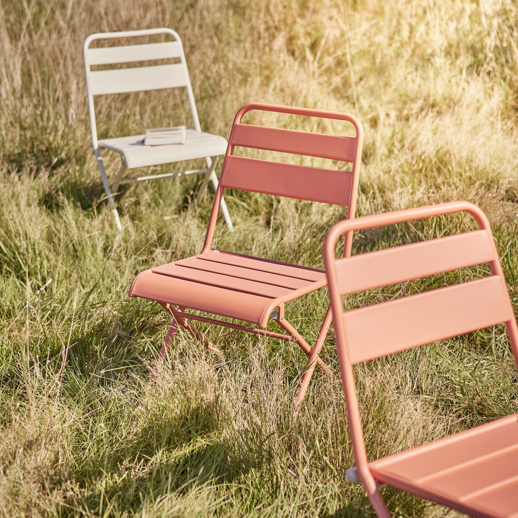 Drei farbenfrohe Gartenstühle aus Metall in Orange und Weiß auf einer sonnigen Wiese im modernen Bistro-Stil.