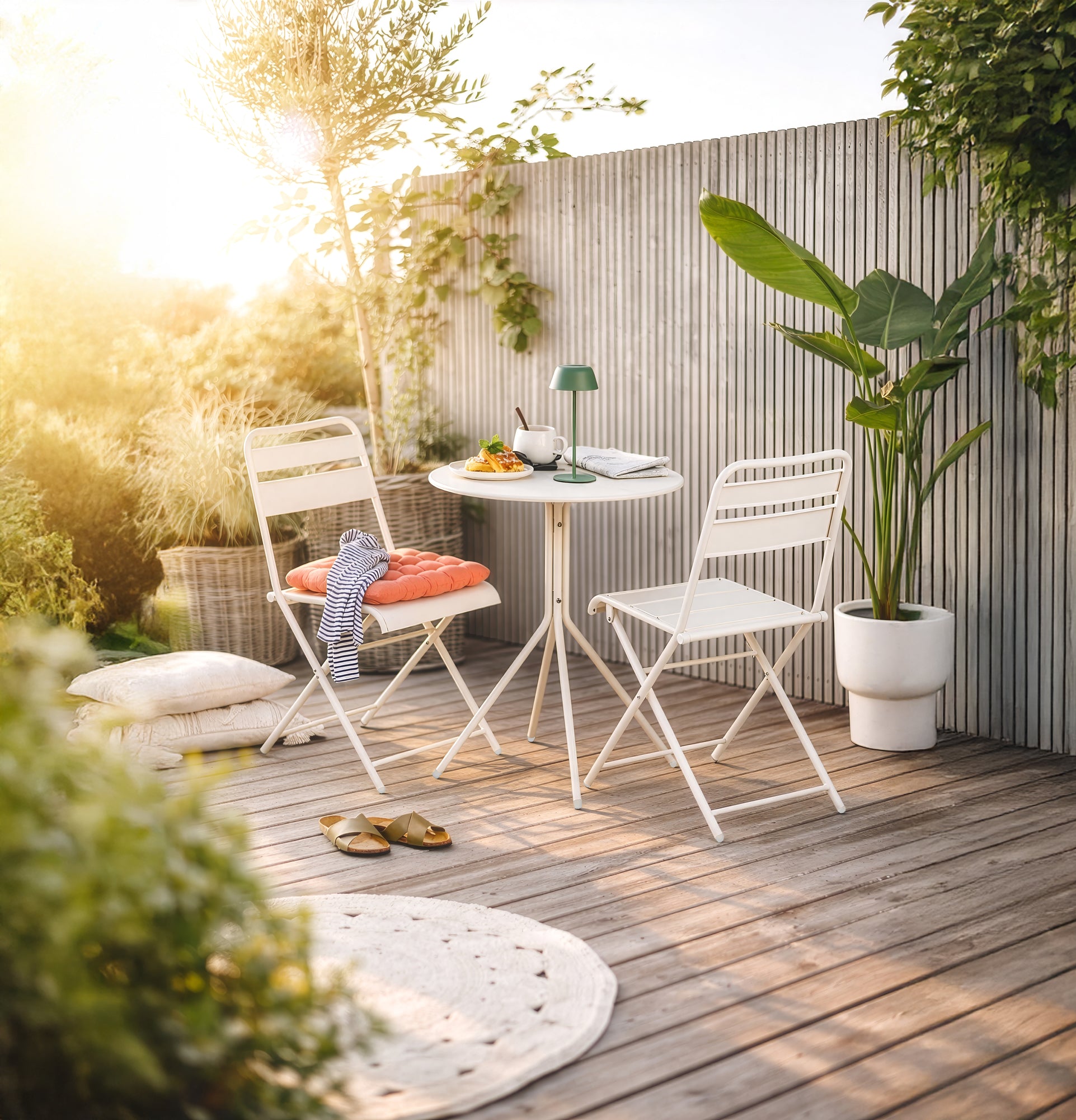 Weißes Gartenmöbel Set aus Metall mit einem runden Bistrotisch und klappbaren Stühlen auf einer Holzterrasse bei Sonnenuntergang.