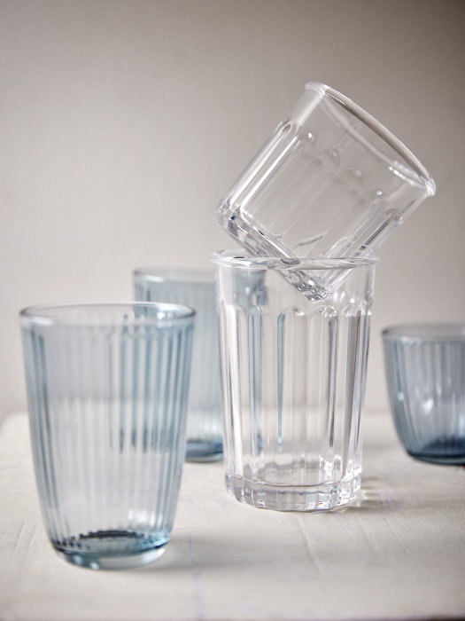 Fünf moderne Wassergläser in verschiedenen Größen und Formen, von geriffelter Struktur bis hin zum klassischen Zylinder-Design, stilvoll auf einem Tisch vor einer weißen Wand arrangiert.